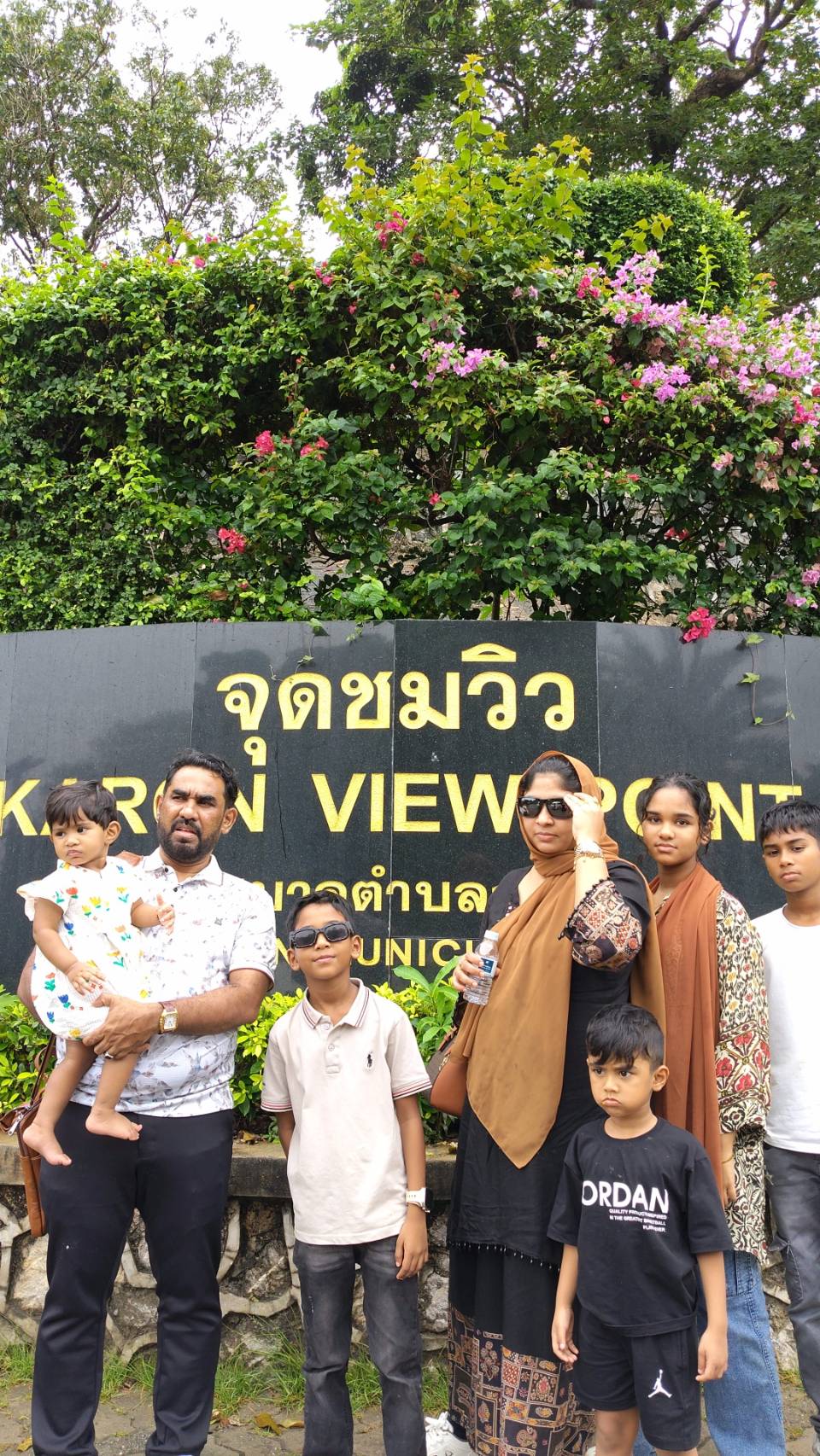 Family posing by the Karon Viewpoint sign amid flowering greenery in Phuket